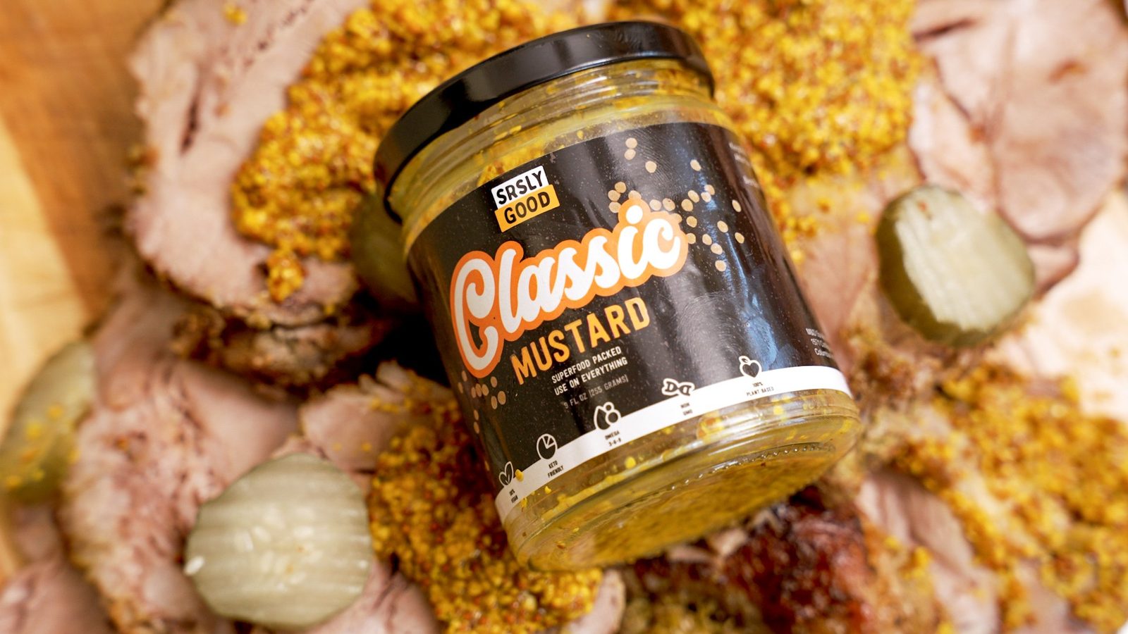 A jar of SRSLY GOOD Classic Mustard on a board of sliced pork, pickles and whole-grain mustard