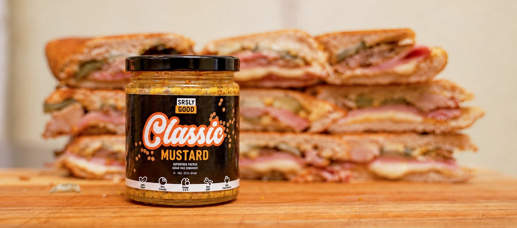 A jar of SRSLY GOOD Classic Mustard on a wooden board in front of stacks of pressed Cuban sandwiches