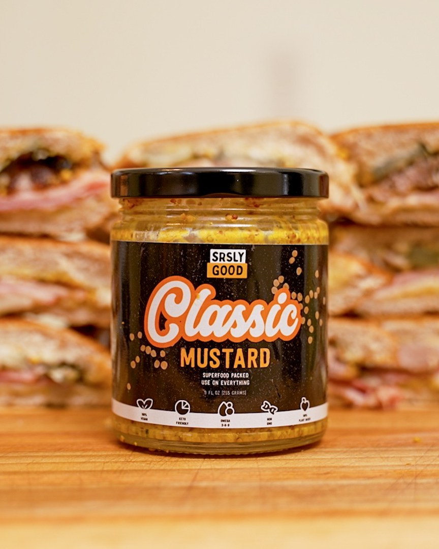 A jar of SRSLY GOOD Classic Mustard on a wooden board in front of stacks of pressed Cuban sandwiches