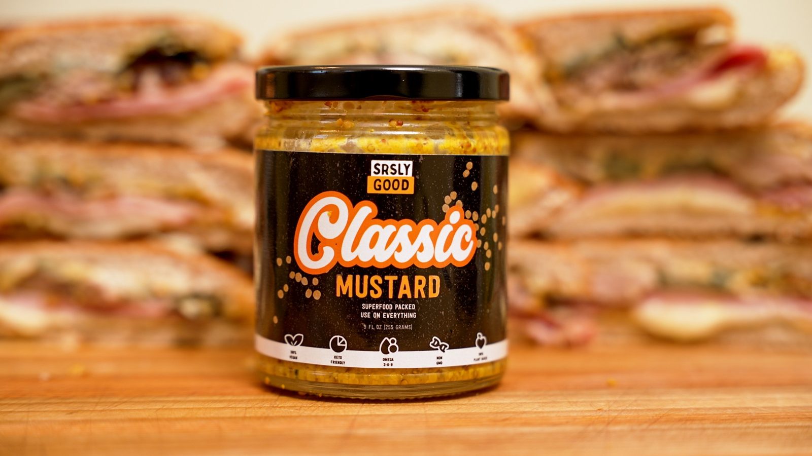 A jar of SRSLY GOOD Classic Mustard in front of a stack of pressed sandwiches